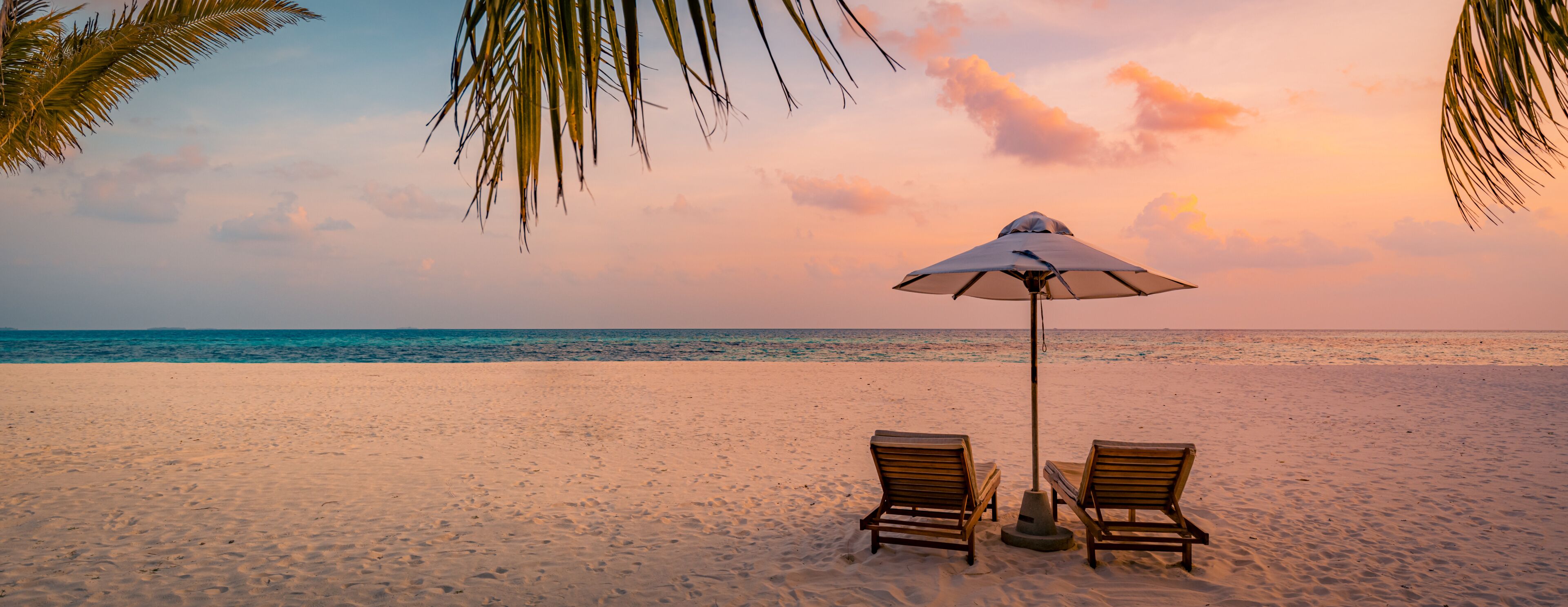 A beach with two wooden lounge chairs and a single umbrella under a palm tree.