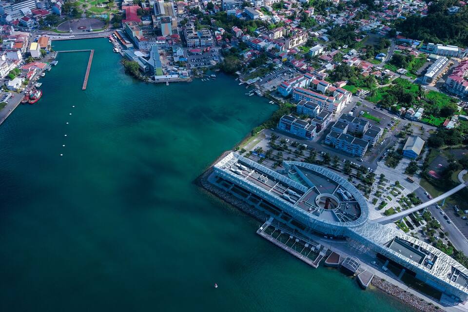 Une vue aérienne d’une ville côtière, avec une promenade en bord de mer bien visible, un grand bâtiment circulaire doté d’un toit particulier et des quartiers résidentiels urbains densément peuplés.