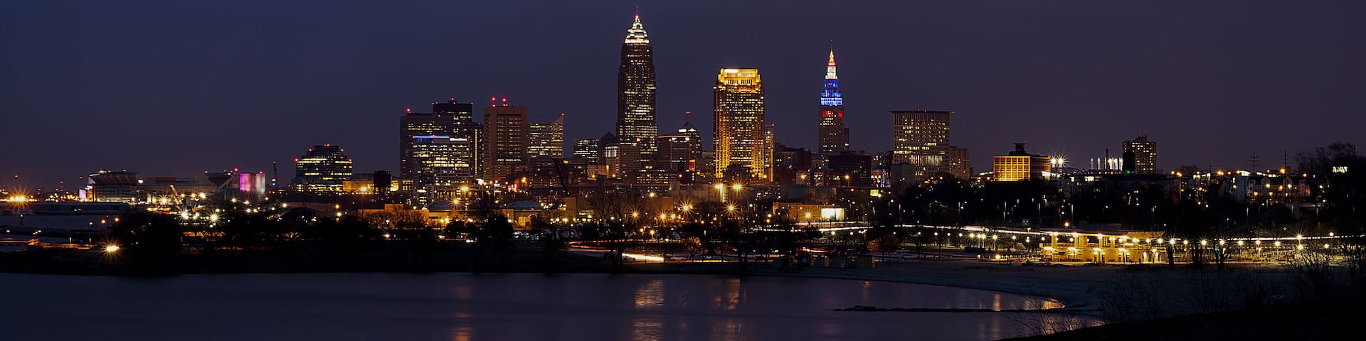 A nighttime city skyline with illuminated buildings and a body of water in the foreground.