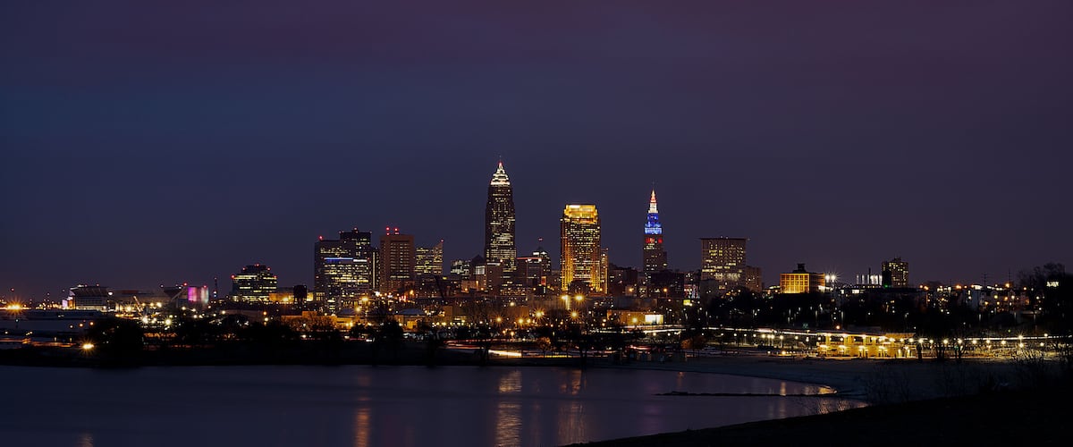 A nighttime city skyline with illuminated buildings and a body of water in the foreground.