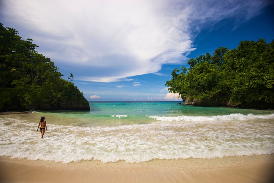 A woman walking on a sandy beach with turquoise water and lush green foliage.