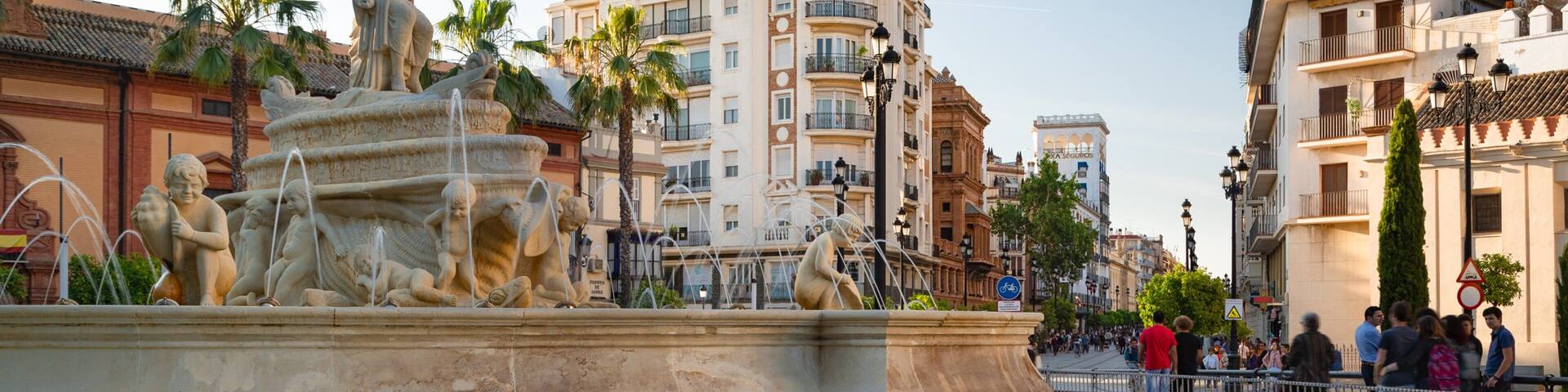 A classical fountain with statues, surrounded by a well-maintained lawn and urban buildings.