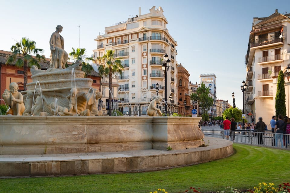 Una fuente clásica con estatuas, rodeada de un césped cuidado y edificios urbanos.