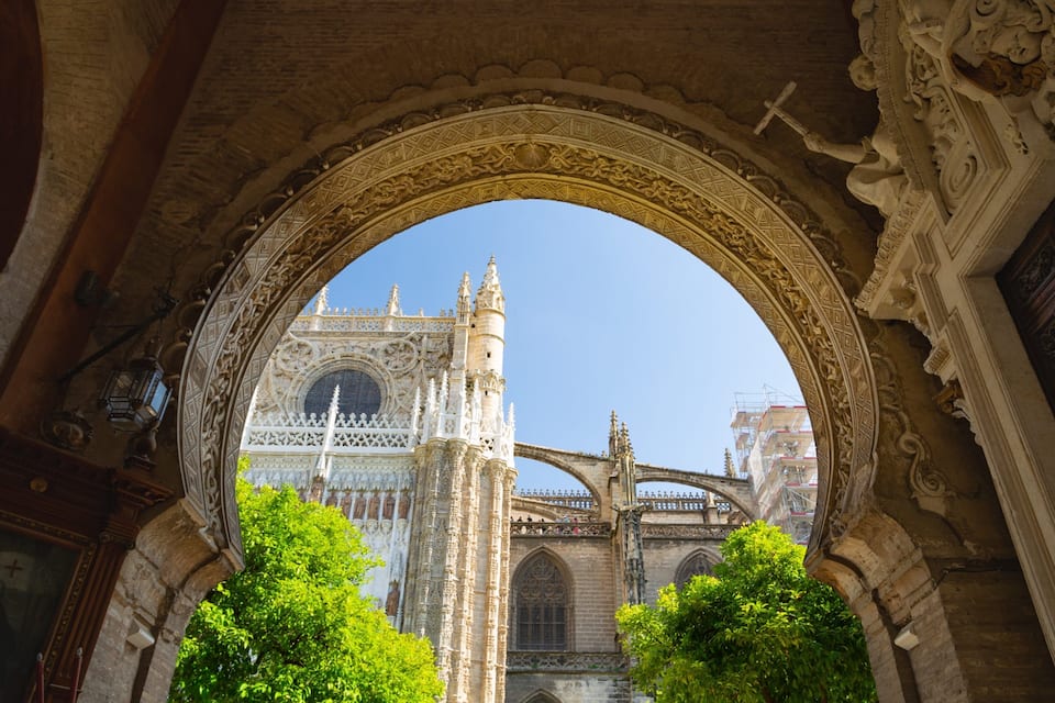 An ornate stone archway framing a view of a historic cathedral with a large rose window and a bridge in the background.