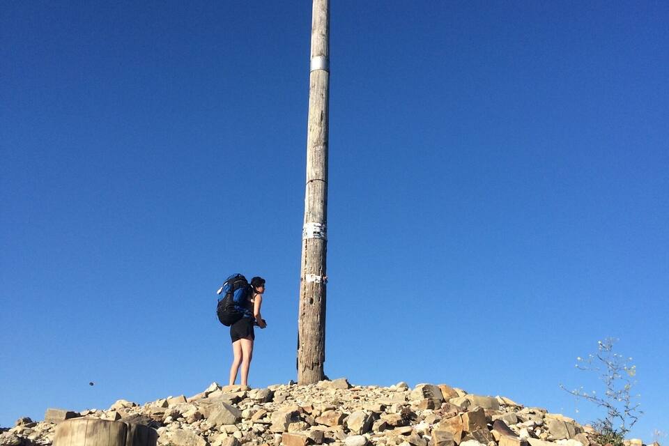 Uma pessoa em uma colina rochosa, com um poste de madeira alto e uma cruz no topo.