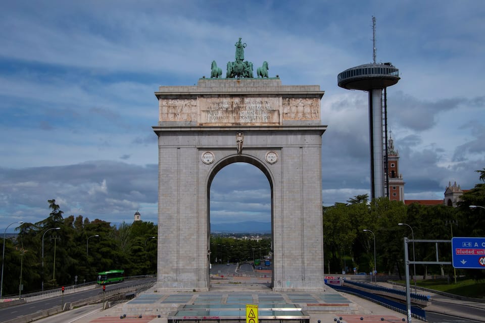 Un gran arco de piedra con una estatua en la parte superior, una torre y una carretera con un autobús verde.