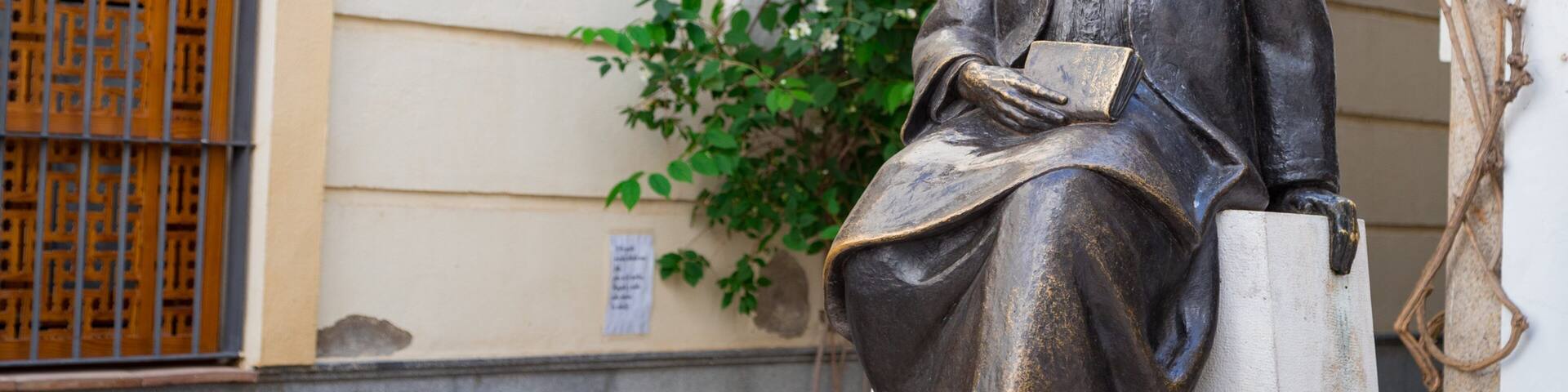 Una estatua de un hombre sentado con un libro, frente a un edificio con una ventana y follaje.