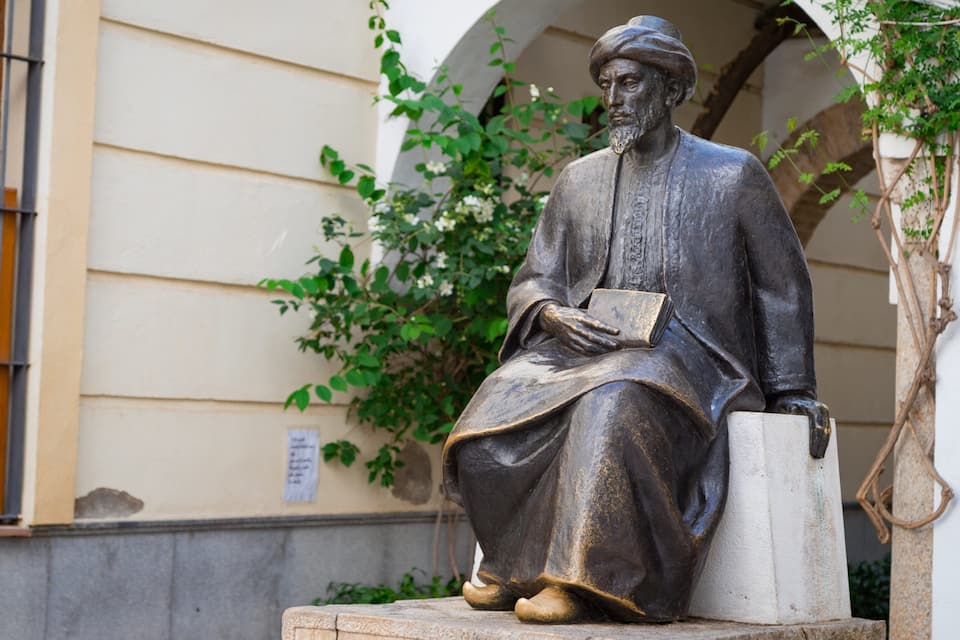 Une statue représentant un homme assis, tenant un livre, placée devant un bâtiment doté d’une fenêtre et agrémenté de verdure.