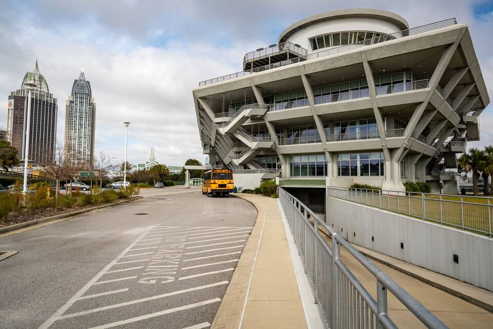 A modern building with a unique angular design, a bus in the foreground, and a cityscape with tall buildings in the background.
