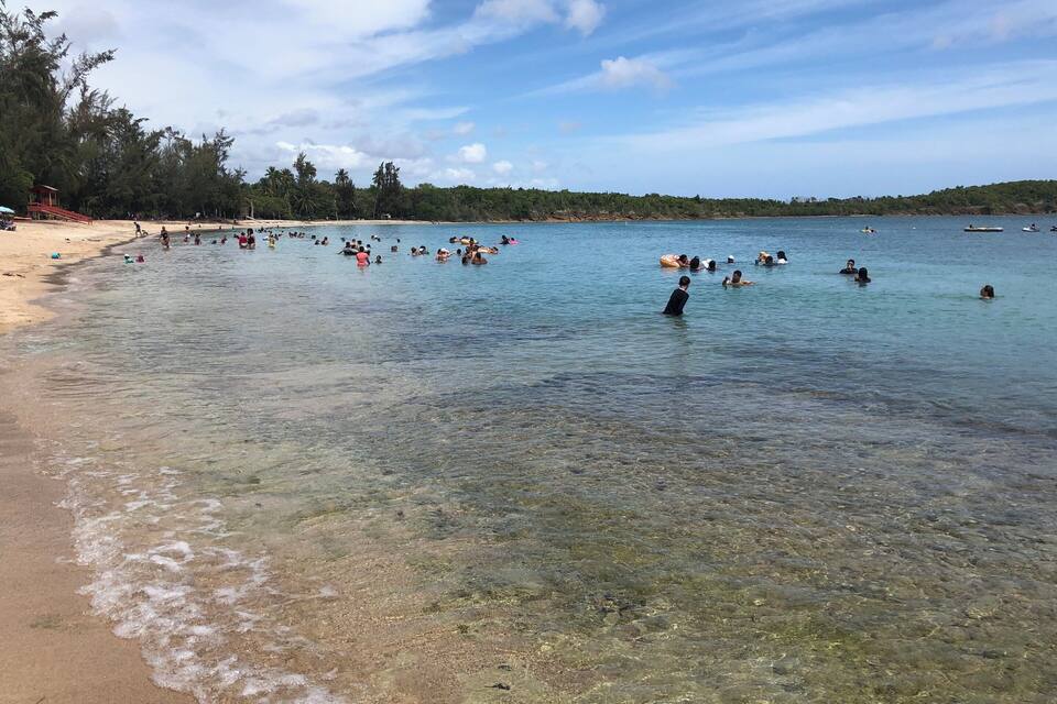 A beach with clear shallow waters and people swimming and wading.
