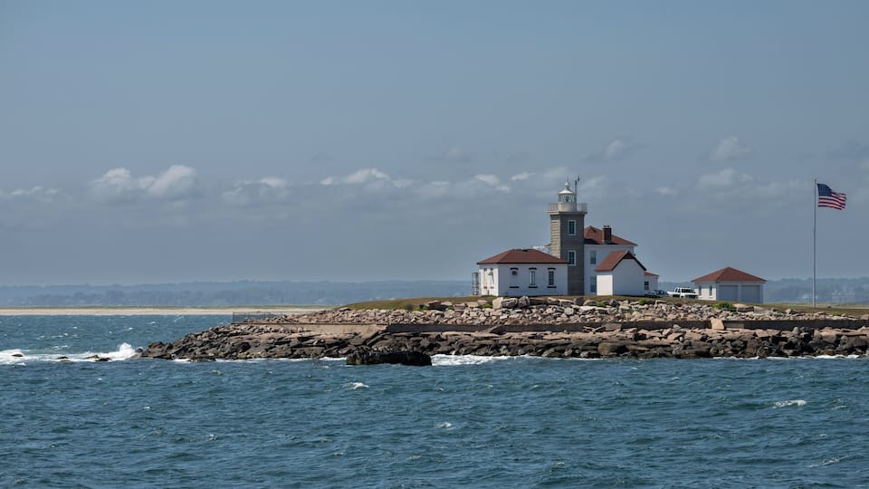 A lighthouse on a rocky outcrop with a flag, surrounded by calm sea and a clear sky.