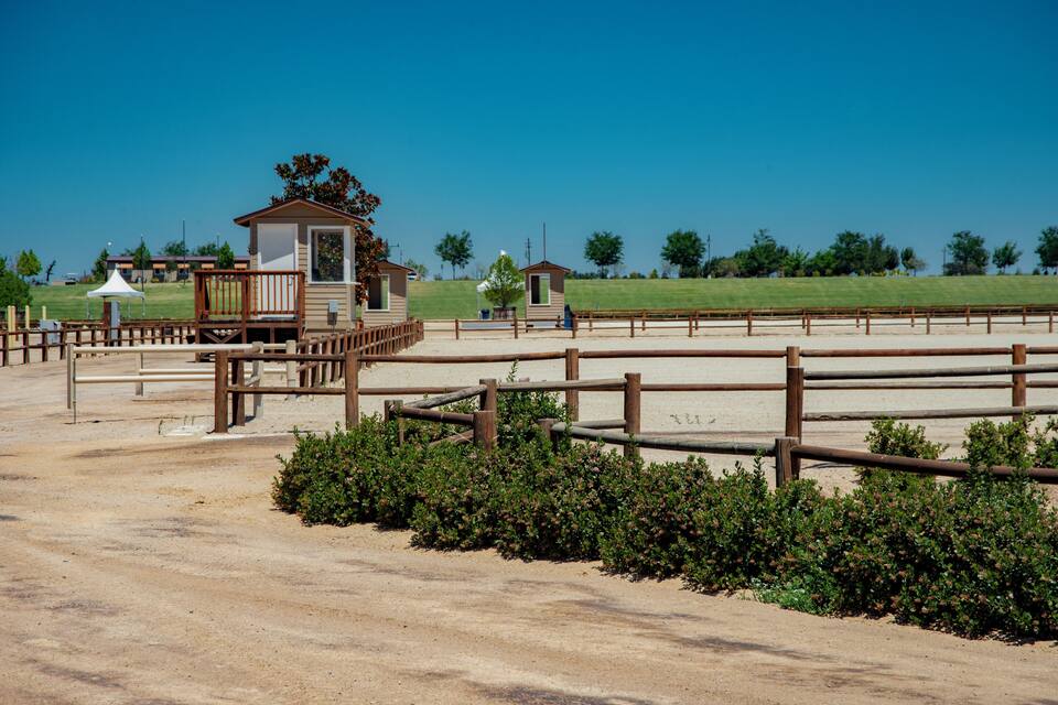 Uma arena equestre cercada por vegetação, com um pequeno edifício de madeira e céu azul claro.
