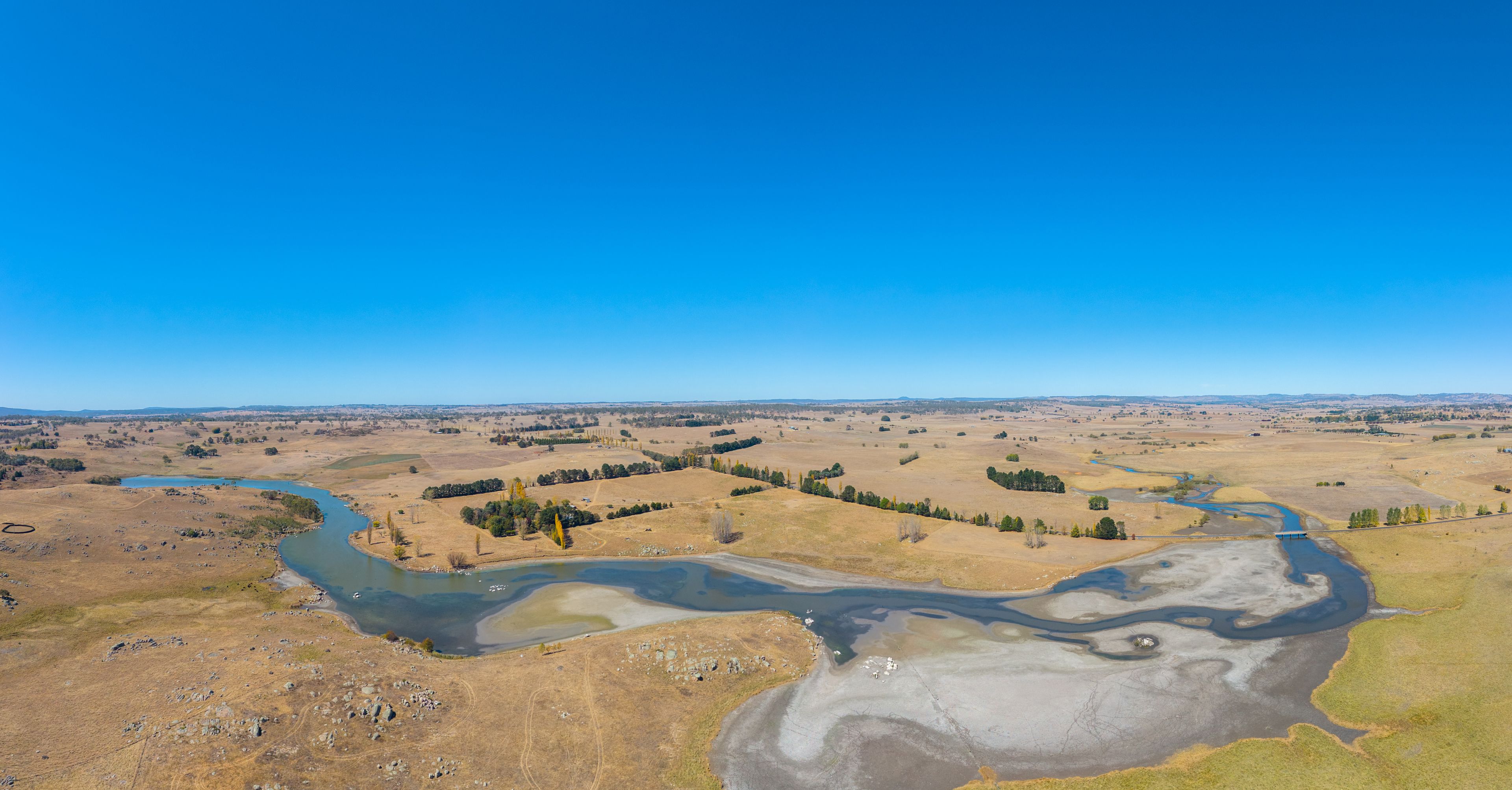 Une rivière serpente à travers un paysage aride et dégagé, parsemé de quelques arbres et bordé de collines lointaines.