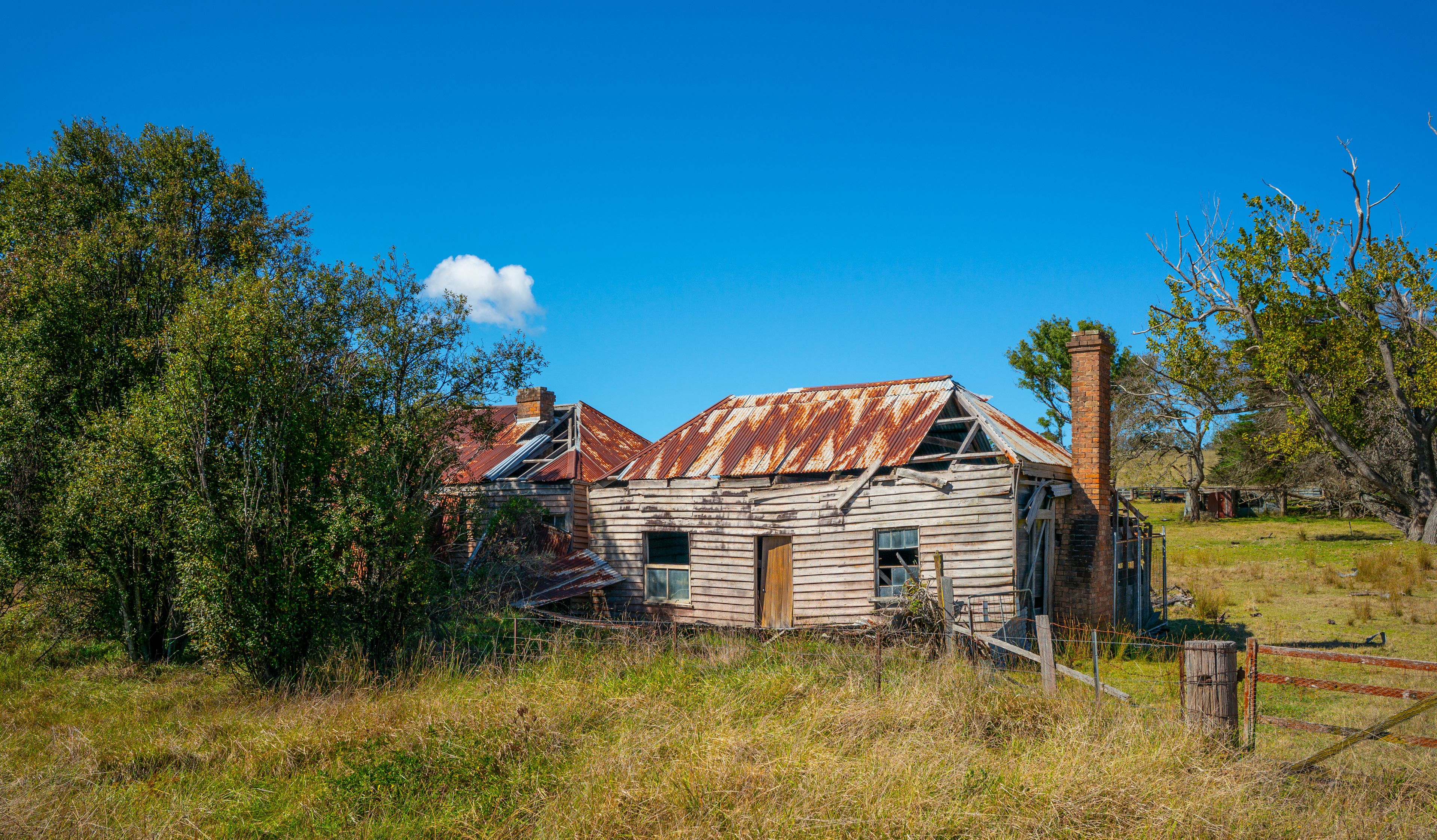 Une vieille maison en bois, dont le toit est rouillé, entourée d’arbres et d’herbe.