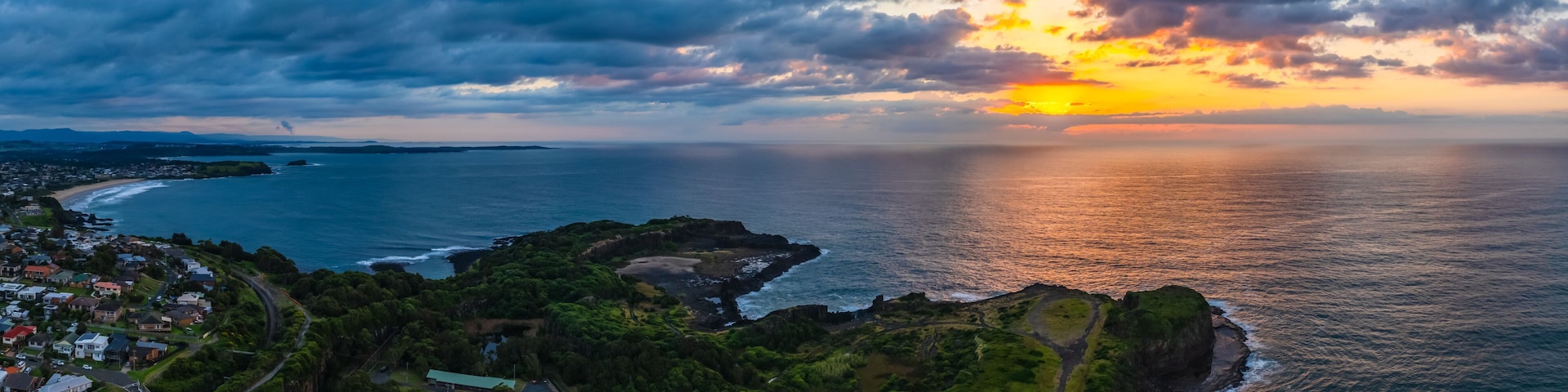 A coastal landscape with a sunset, a rocky coastline, and a residential area.