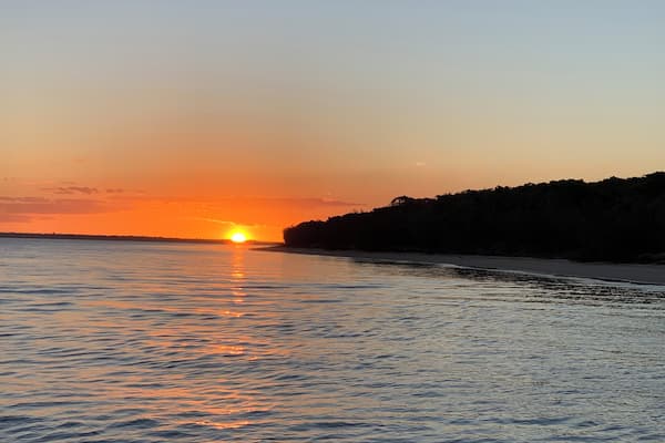 A sunset over a calm sea with a beach and a forested hill in the background.