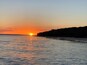 A sunset over a calm sea with a beach and a forested hill in the background.