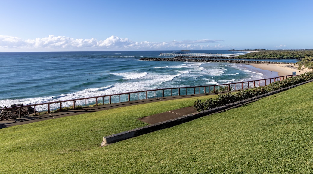 A coastal landscape with a grassy area, a beach, waves, a pier, and a clear sky.
