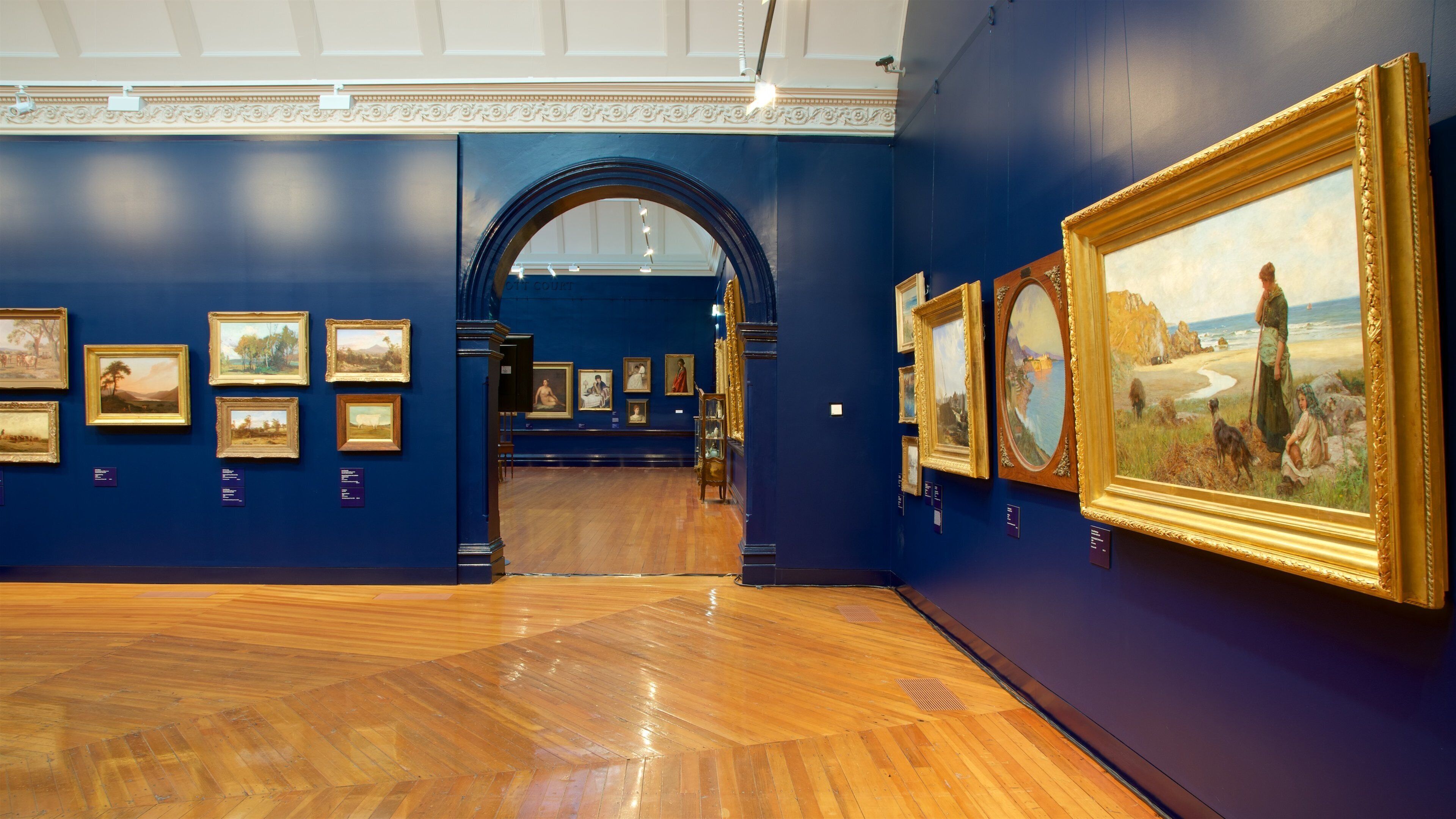 An art gallery with wooden floors and blue walls, featuring various framed paintings.
