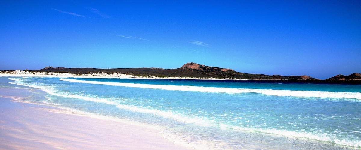 A beach with clear turquoise water, white sand, and distant mountains under a blue sky.
