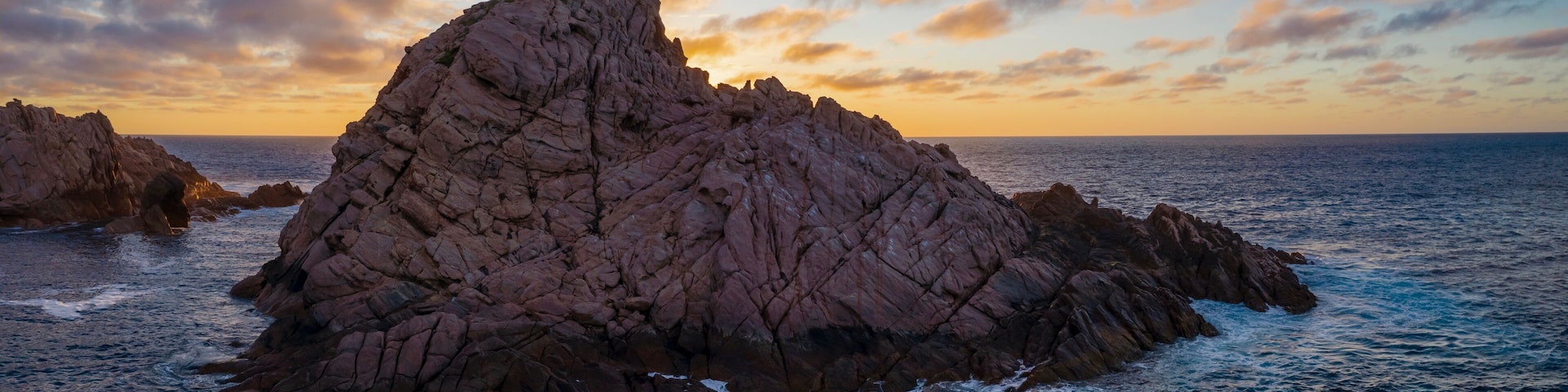 A rugged rock formation surrounded by ocean waves at sunset.