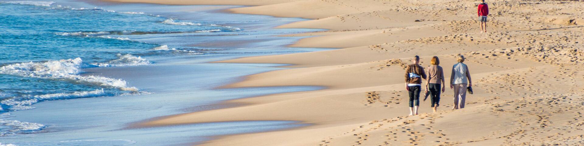 A sandy beach with gentle waves, footprints, and a person in a red jacket walking away.