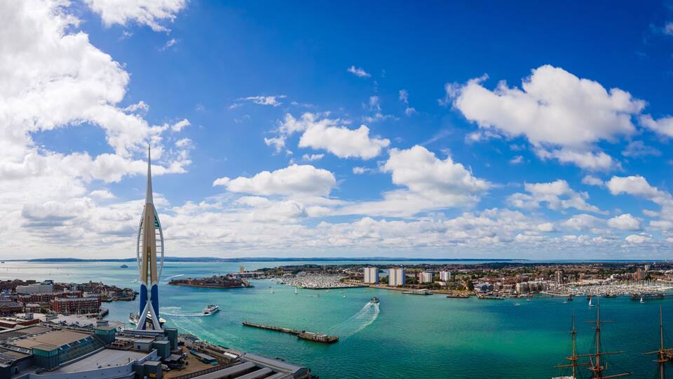 A coastal cityscape with a prominent tower, a marina with boats, and a clear blue sky.