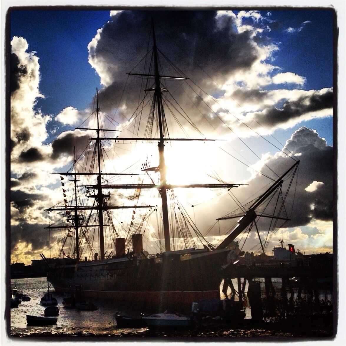 Et stort sejlskib ligger til ankers i en havn under en delvist overskyet himmel.