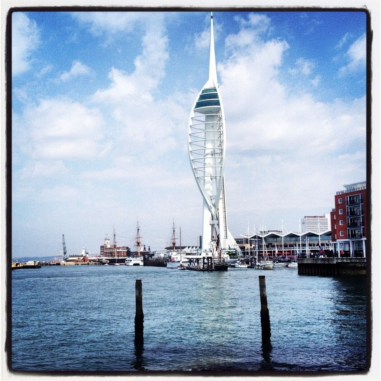 A modern sail-shaped building by the water with boats and construction cranes in the background.