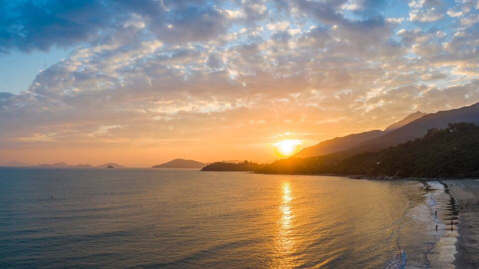 A beach with a sunset over the ocean, mountains in the background, and a clear sky.