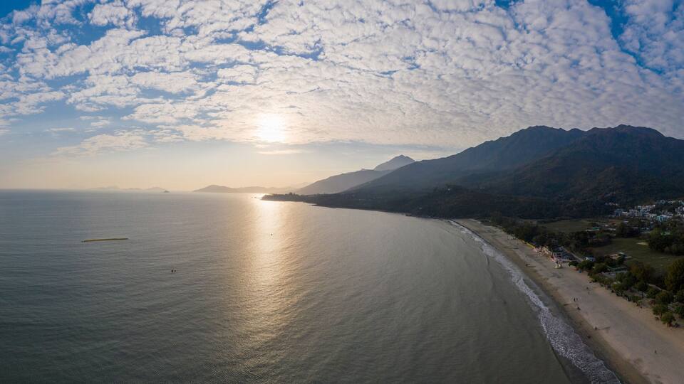 A coastal landscape with a beach, mountains, and a body of water reflecting the sun.