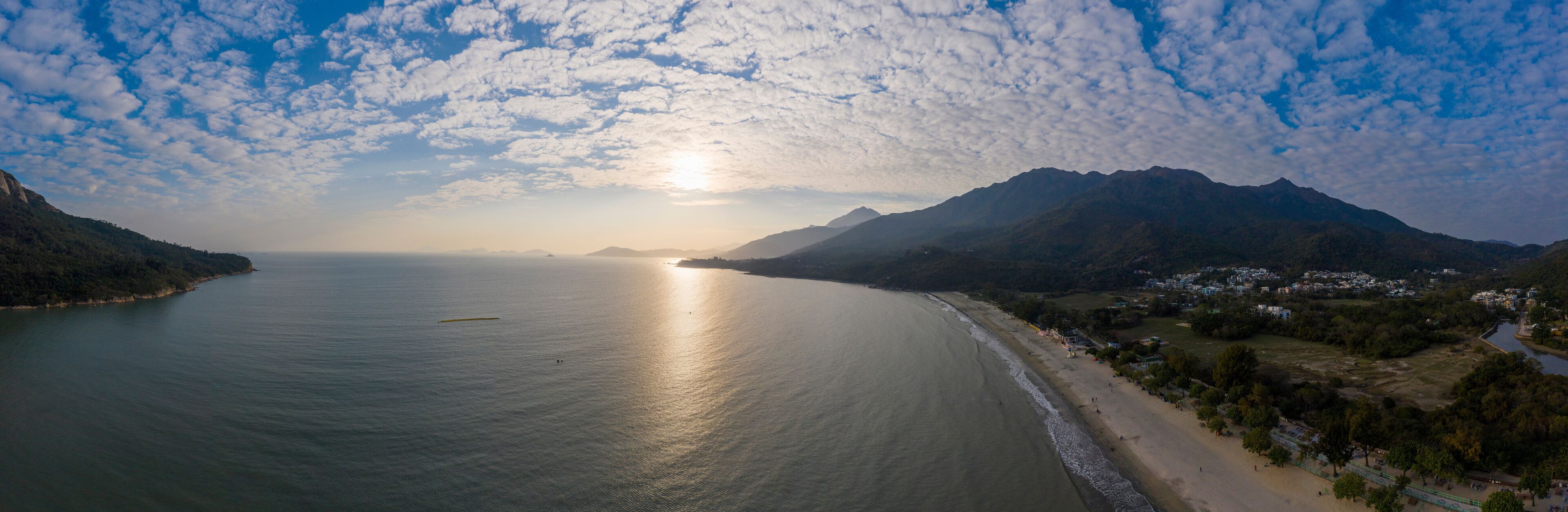 A coastal landscape with a beach, mountains, and a body of water reflecting the sun.