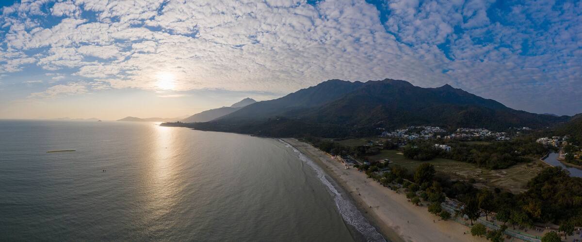 A coastal landscape with a beach, mountains, and a body of water reflecting the sun.