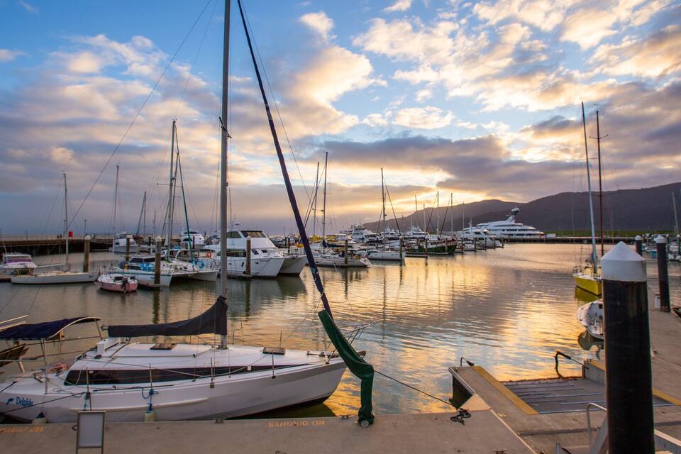 Un puerto deportivo con varias embarcaciones amarradas, cielo despejado y montañas al fondo.