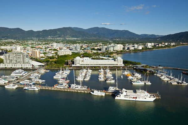 A marina with numerous boats docked, a large cruise ship, and a cityscape with buildings and mountains in the background.
