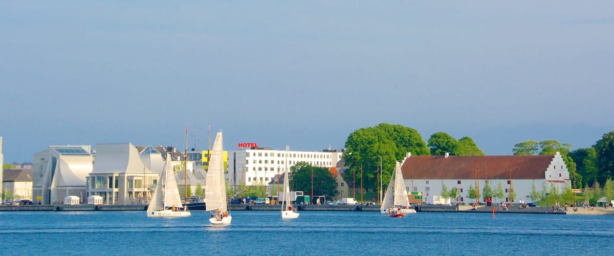 Sailboats on a calm river, a modern building with a 'HOTEL' sign, and a traditional building with a red roof.