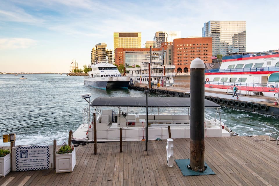 A waterfront scene with a boat docked at a pier, a city skyline in the background, and a red and white ferry on the right.