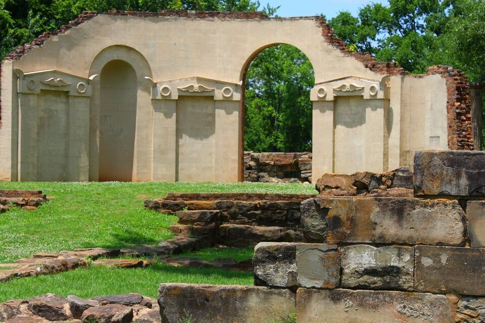 An ancient stone structure with arches and columns, surrounded by grass and trees.