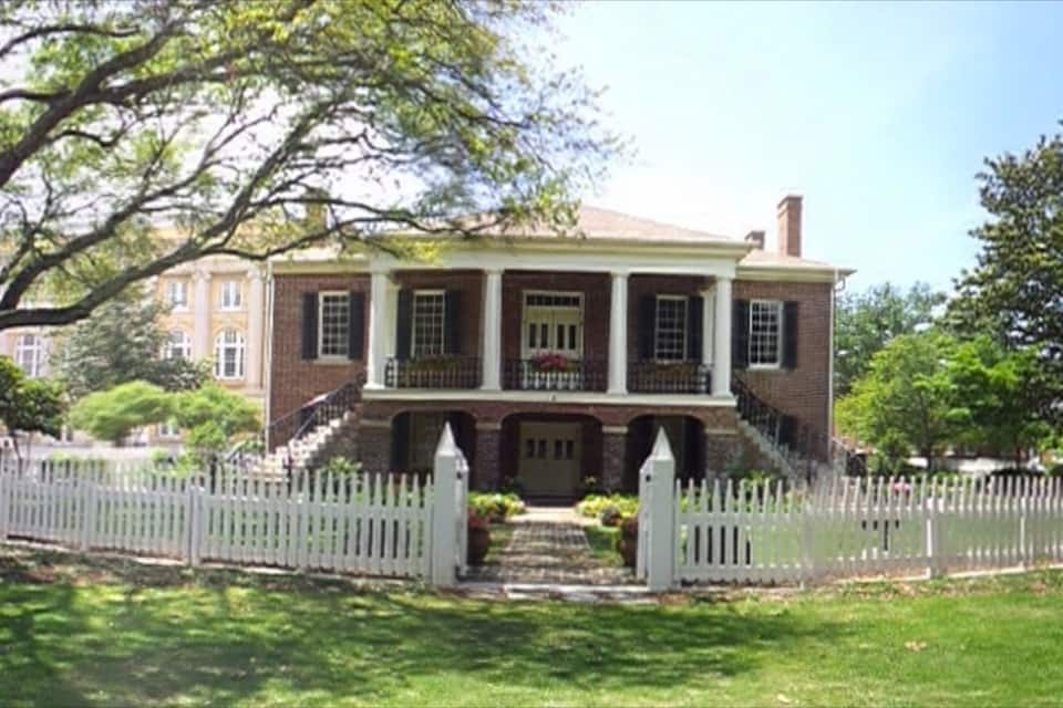 A two-story brick house with a white picket fence, surrounded by lush greenery and trees.