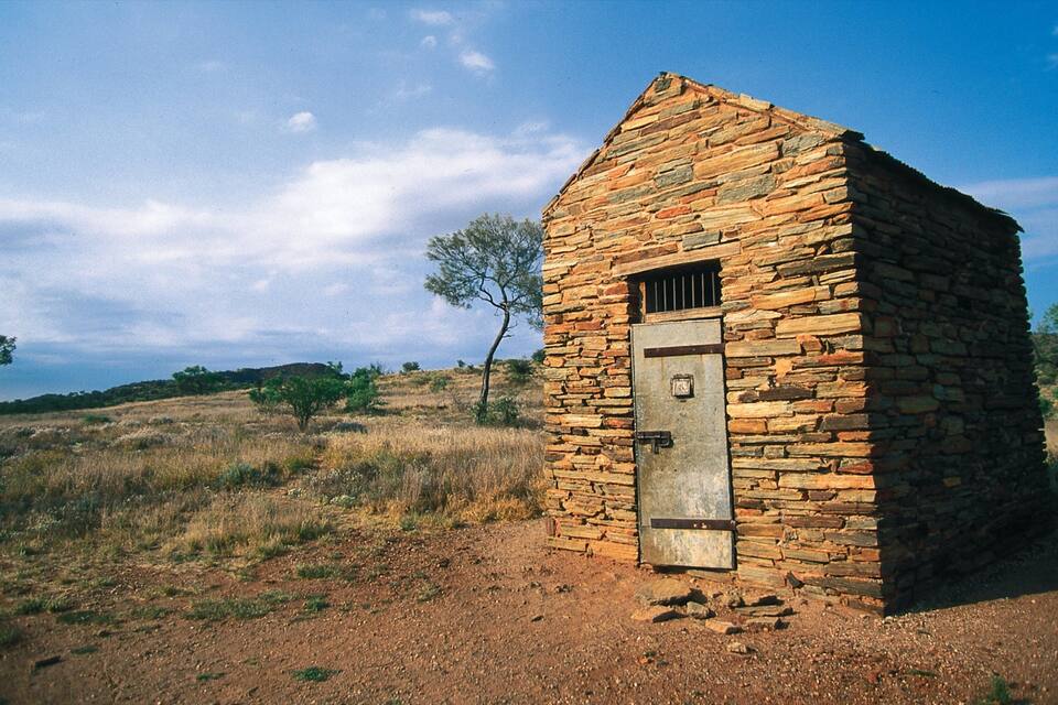 A small stone building with a metal door in a dry, grassy landscape with sparse trees.