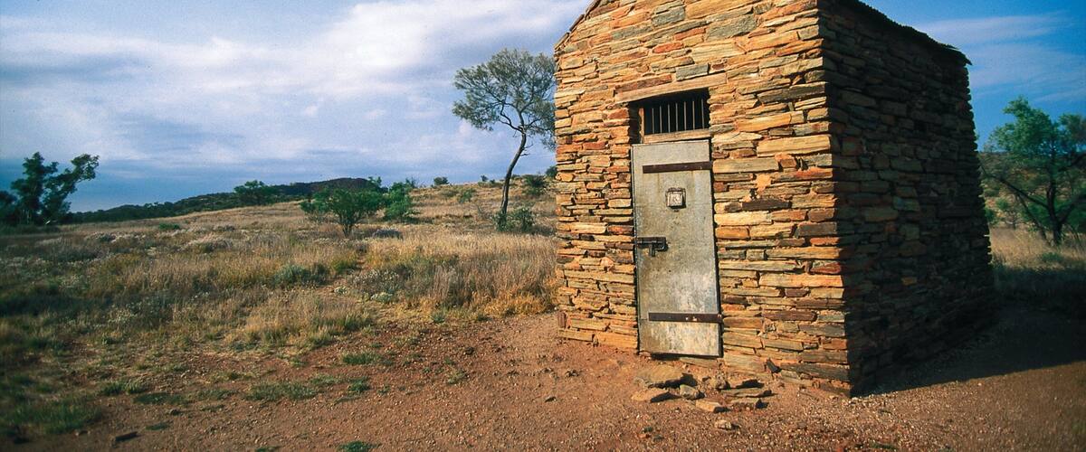 A small stone building with a metal door in a dry, grassy landscape with sparse trees.