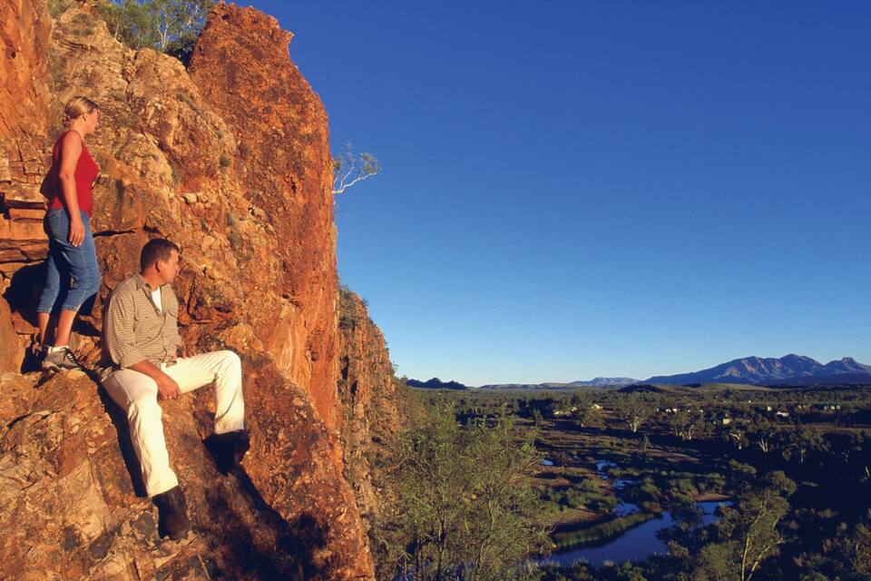 Un hombre y una mujer sentados en un acantilado rocoso rojo, con vistas a un valle con un río y montañas a lo lejos.