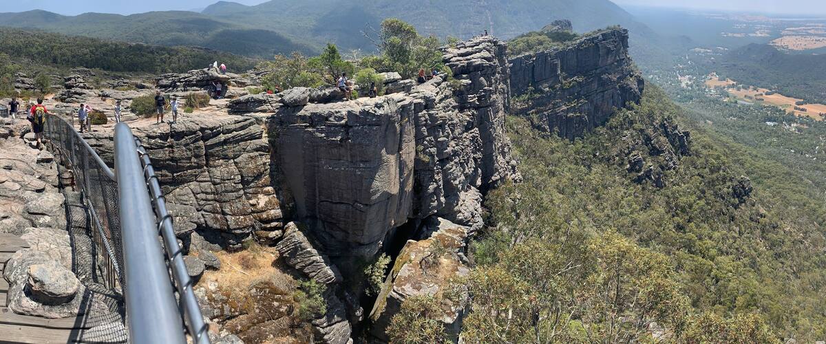 A rocky cliff with a metal railing, surrounded by dense forest and distant mountains.