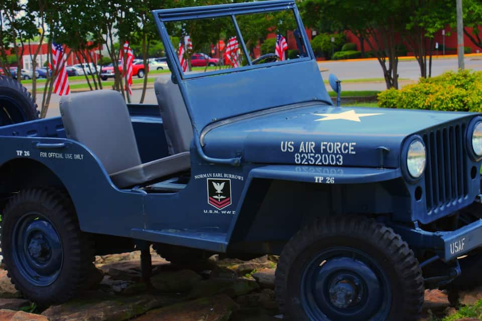 A blue military jeep with US Air Force insignia and a star emblem, parked on a stone surface with American flags in the background.