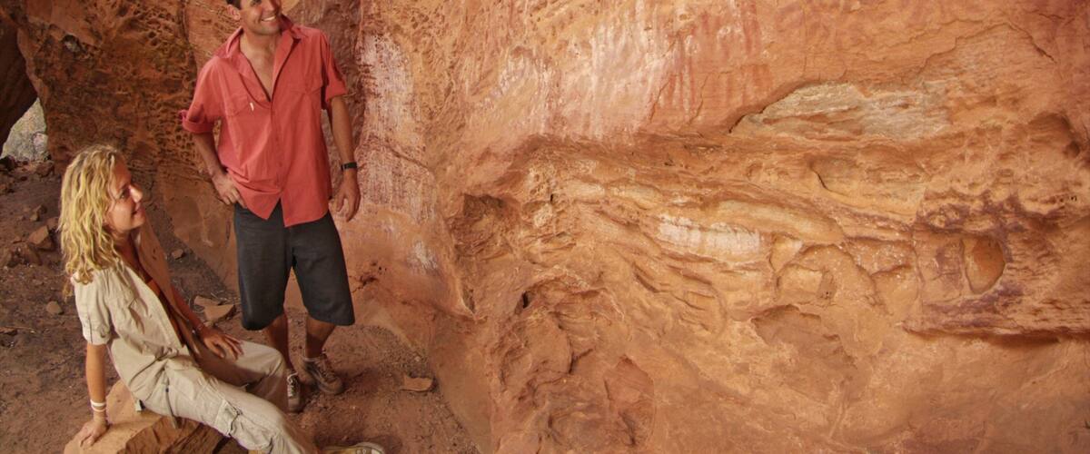 A man in a red shirt and shorts standing in a cave with reddish rock walls.