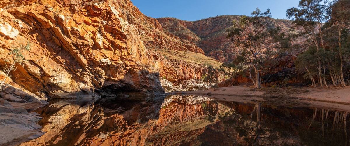 A canyon with red rock formations and a calm river reflecting the scenery.