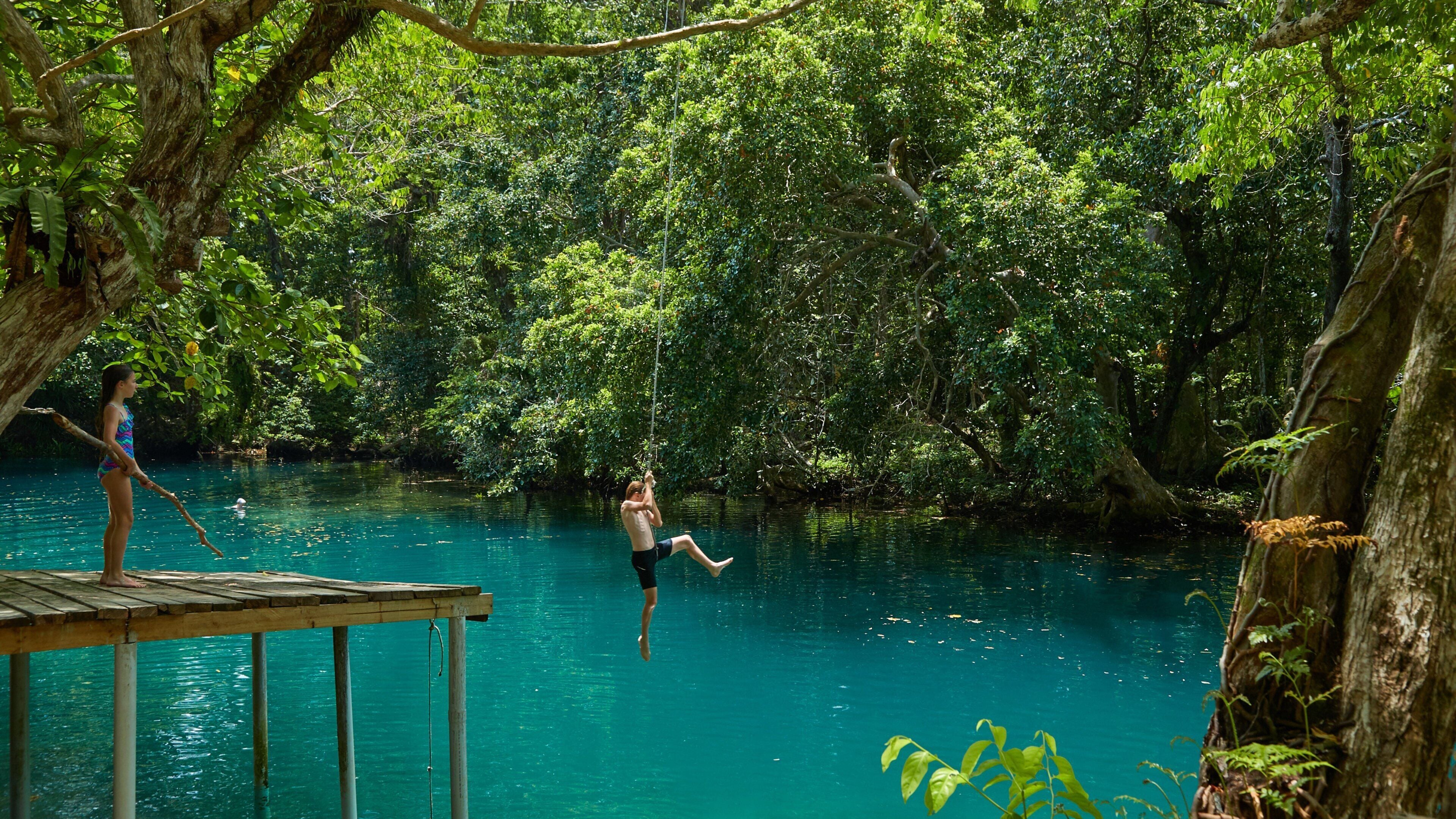 A person swinging on a rope over a turquoise river surrounded by dense green forest.