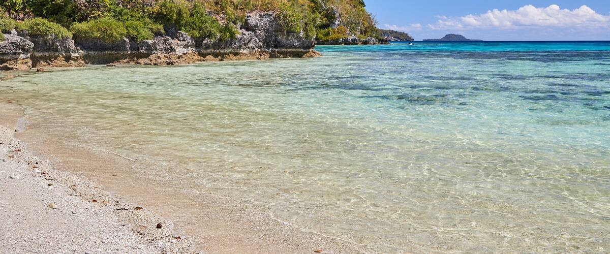 A clear beach with turquoise water, rocky cliffs, and dense green foliage.