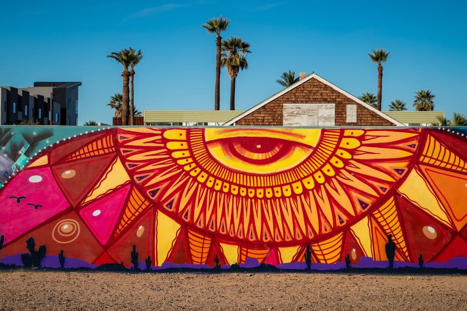 A colorful mural with an eye design, palm trees, and a building in the background.
