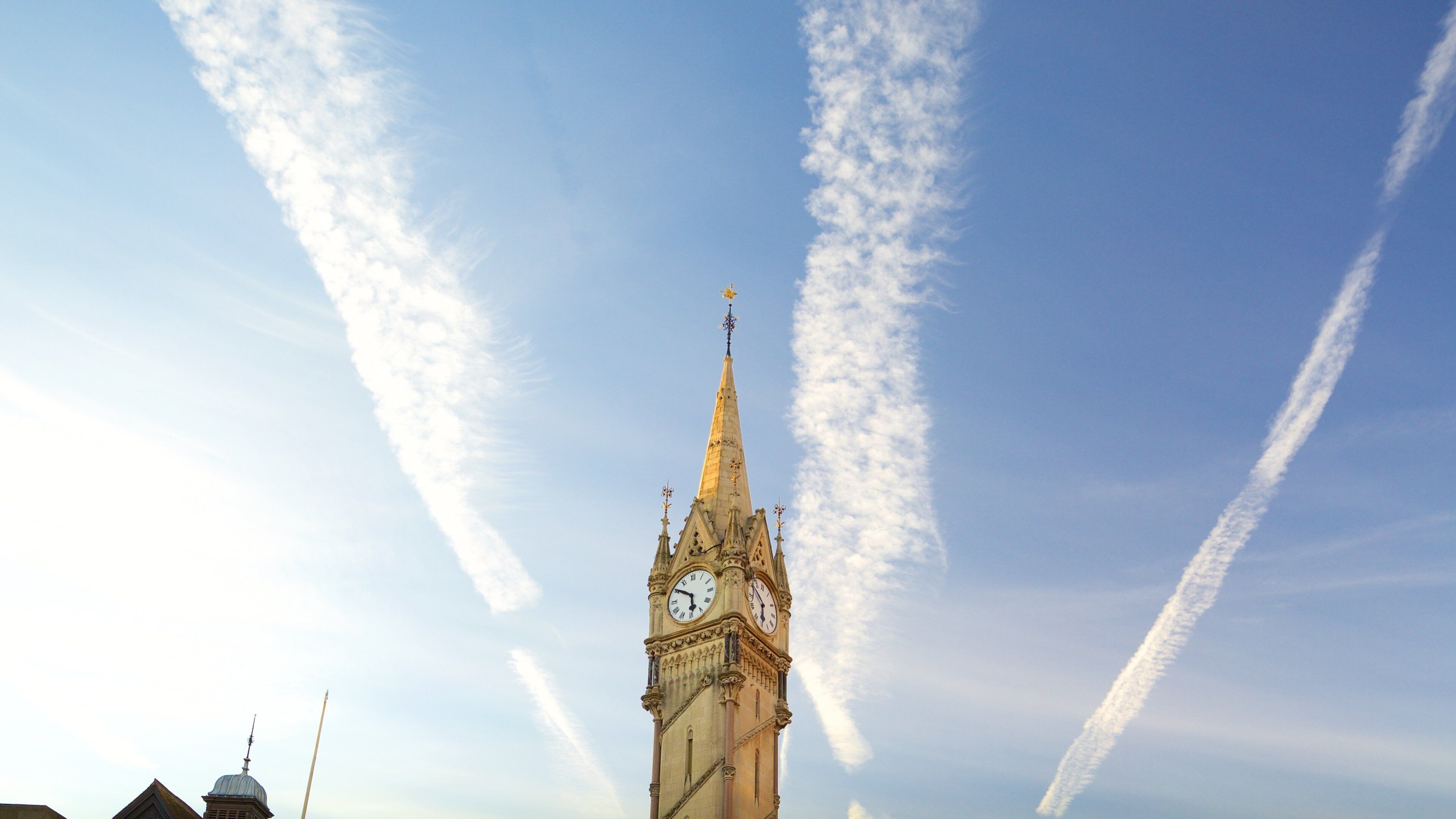 A clock tower with a pointed spire and a weather vane on top, set against a clear sky with contrails.